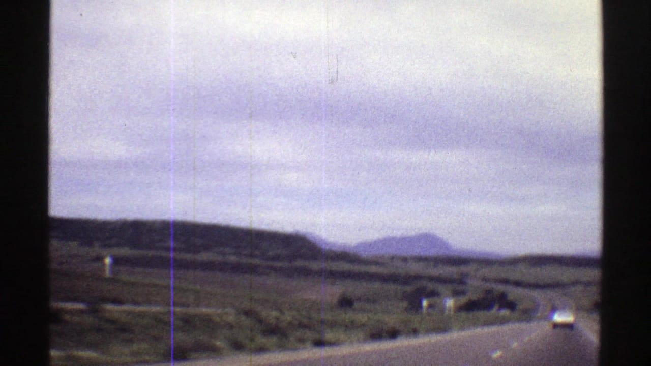 1969 Colorado home-movie frame showing a rural two-lane road, roadside brush, distant hills, and soft film grain.
