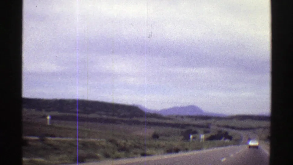 1969 Colorado home-movie frame showing a rural two-lane road, roadside brush, distant hills, and soft film grain.