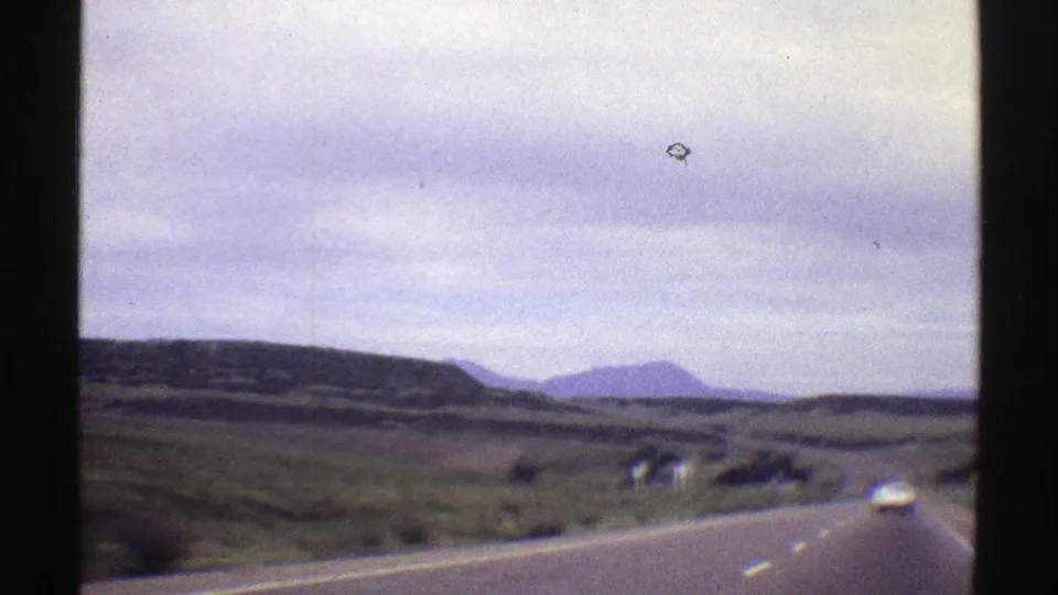 1969 Colorado home-movie view from a car on a rural highway, with open land, distant mountains, and a white car ahead.