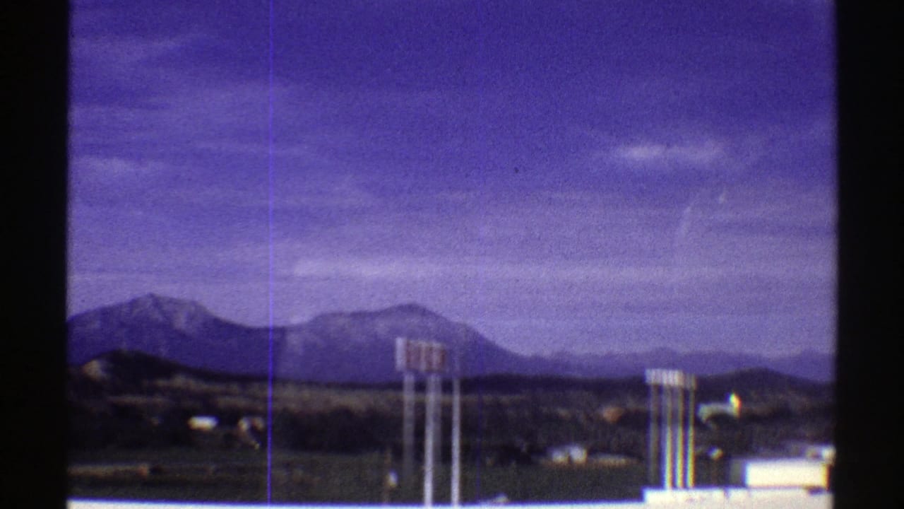 1969 Colorado roadside view with distant mountains, utility poles, low buildings, and a dark tree line under a blue-purple sky.