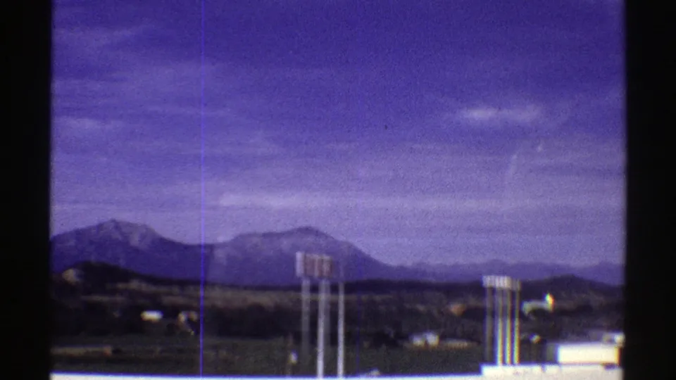 1969 Colorado roadside view with distant mountains, utility poles, low buildings, and a dark tree line under a blue-purple sky.