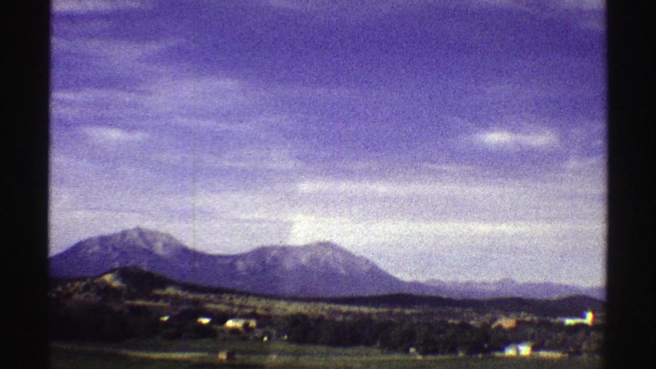 Wide 1969 Colorado home-movie view of mountains beyond open land, scattered buildings, and a clouded sky.