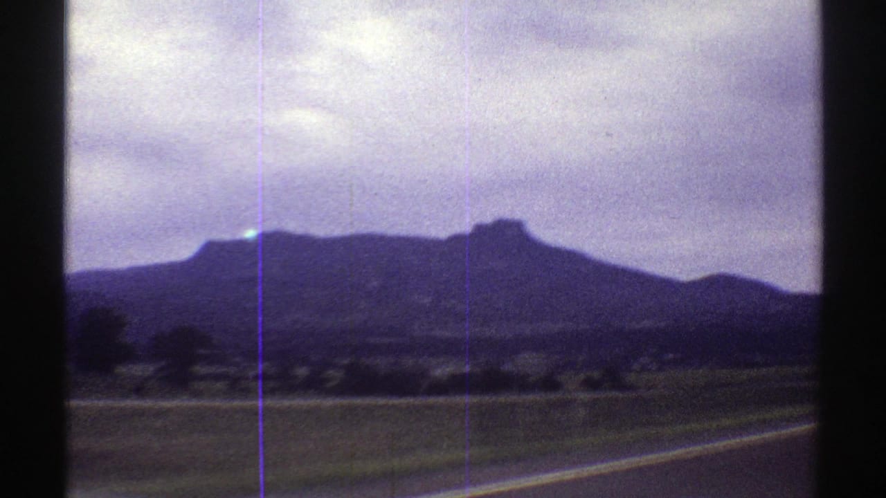 1969 Colorado 8mm frame of a mesa-like landform and roadside fields, shot from a moving car with dashboard visible at the bottom.