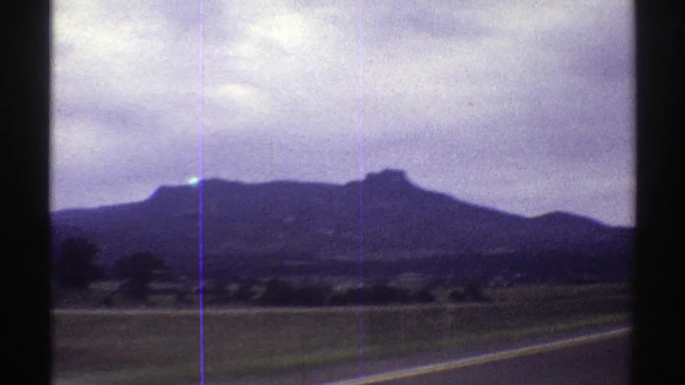 1969 Colorado 8mm frame of a mesa-like landform and roadside fields, shot from a moving car with dashboard visible at the bottom.