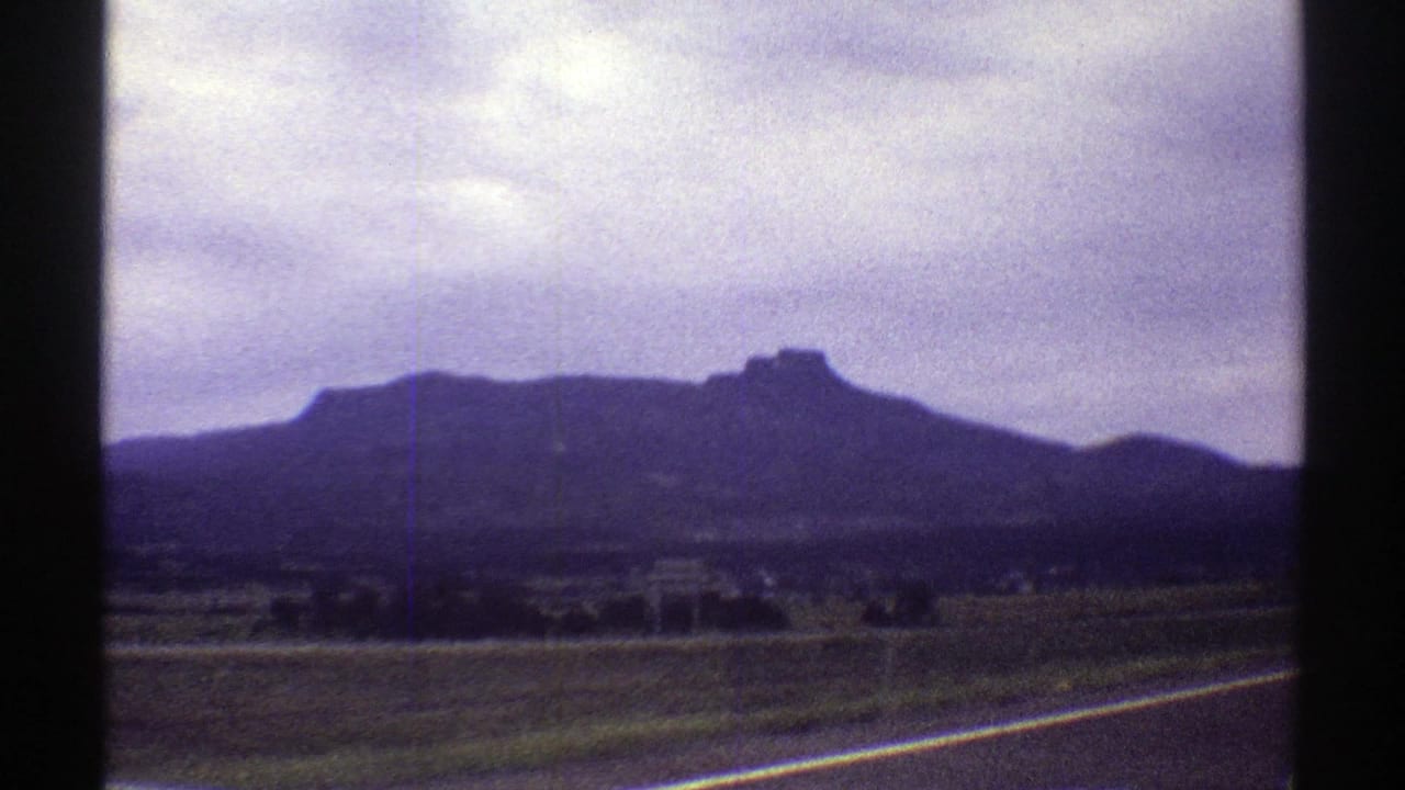 1969 Colorado 8mm frame showing a paved roadside, open fields, scattered trees, and a dark mountain ridge in the distance.
