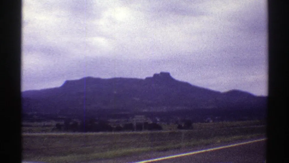 1969 Colorado 8mm frame showing a paved roadside, open fields, scattered trees, and a dark mountain ridge in the distance.