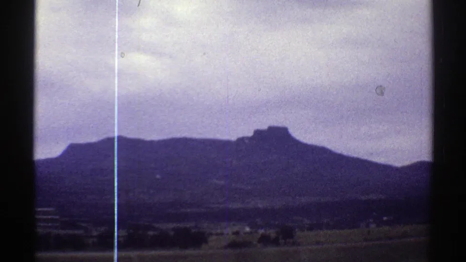 1969 Colorado roadside view with a broad ridge, low fields, faint buildings, cloudy sky, and visible vertical film line.