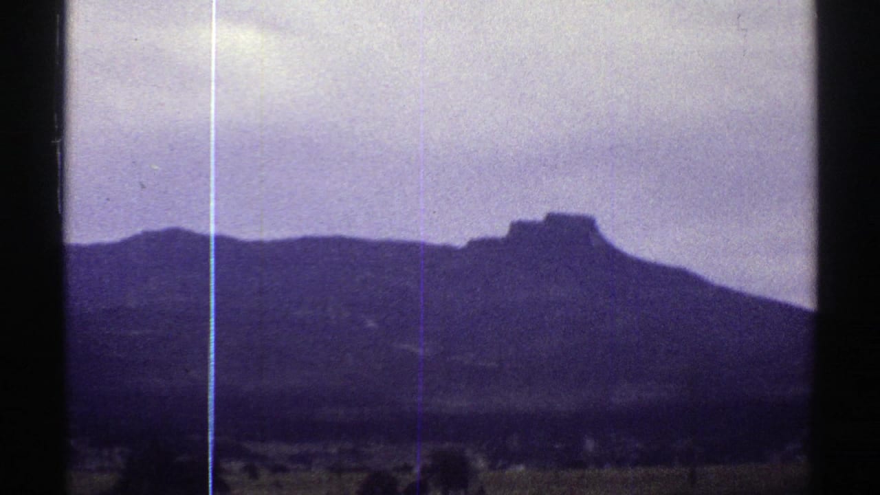 Colorado home-movie frame from 1969 showing open rural ground, distant trees, and a dark mountain profile beneath a pale cloudy sky.