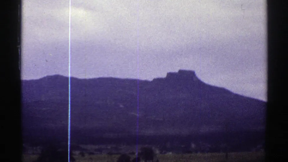 Colorado home-movie frame from 1969 showing open rural ground, distant trees, and a dark mountain profile beneath a pale cloudy sky.