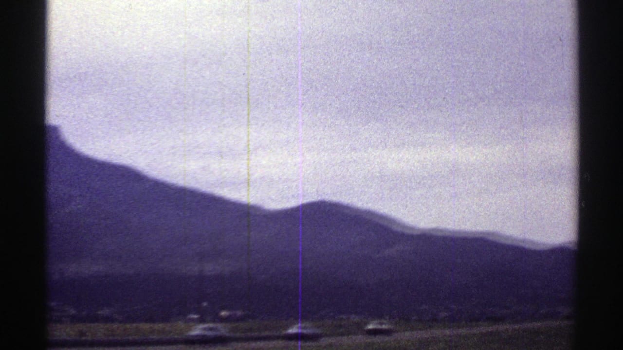 1969 Colorado 8mm view from a moving vehicle, with distant mountains, a rural road edge, cloudy sky, and dark window framing.