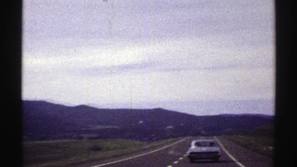 1969 Colorado road scene with a car ahead on a two-lane highway, rolling hills, distant mountains, and overcast sky.