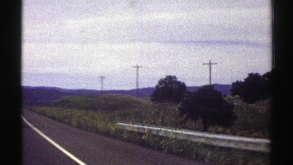 1969 Colorado roadside view from a moving car, showing a guardrail, utility poles, fields, low hills, and cloudy sky.