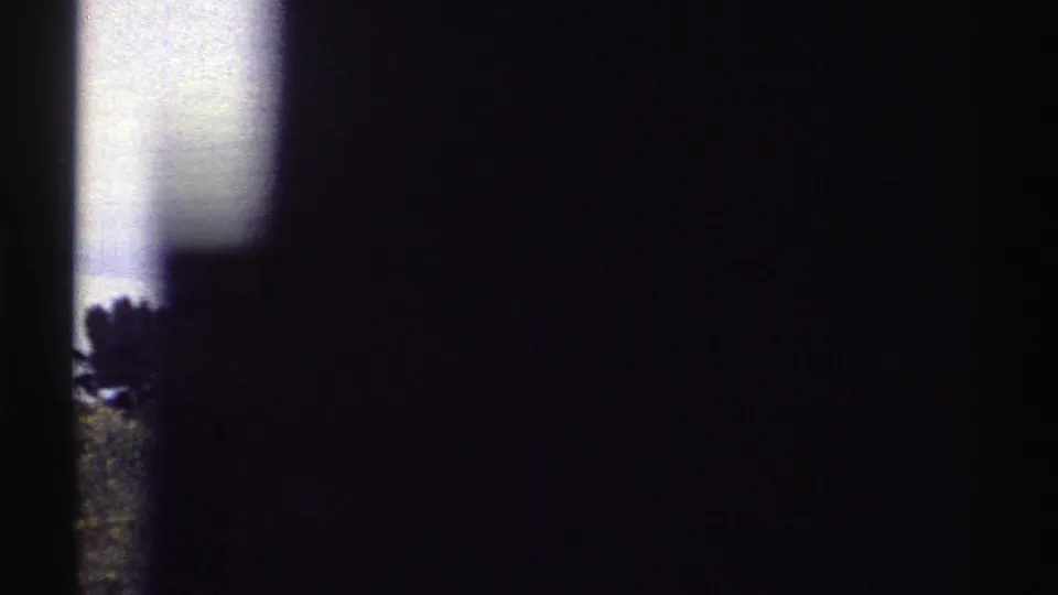 1969 Colorado home-movie frame showing a shadowed vehicle-window view with trees, open ground, and cloudy sky beyond.