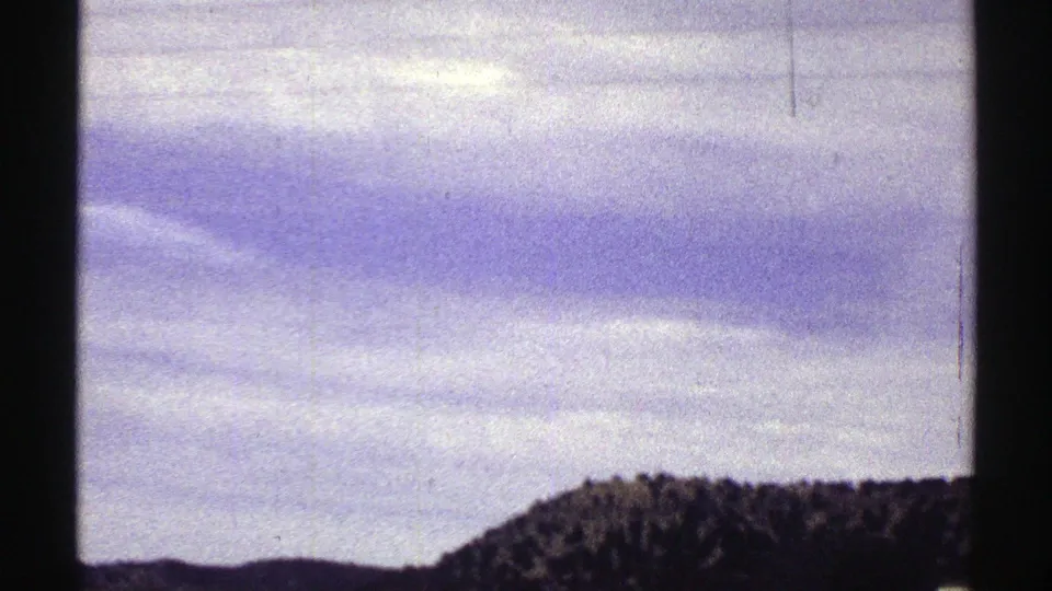 1969 Colorado home-movie frame with a low tree-covered hill, open land, cloudy sky, and dark window framing from inside a vehicle.