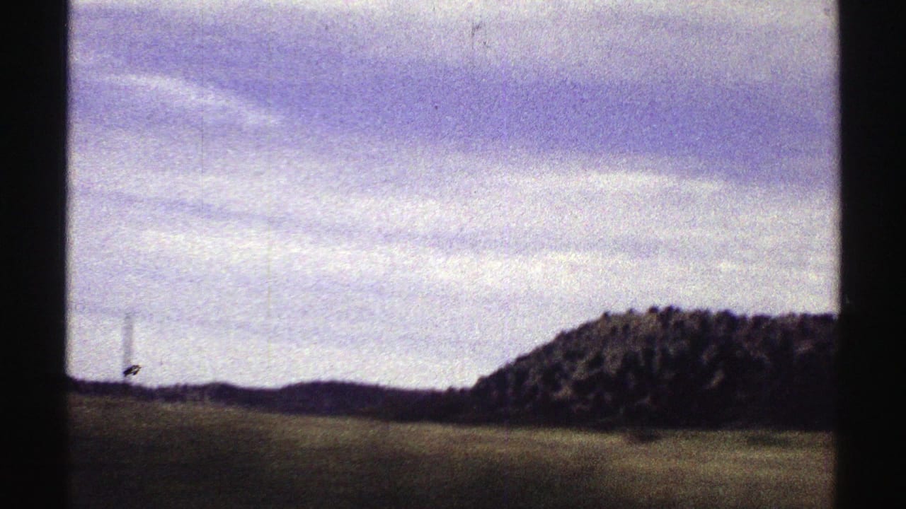 Open field and wooded hill seen through dark vehicle or film-frame edges.