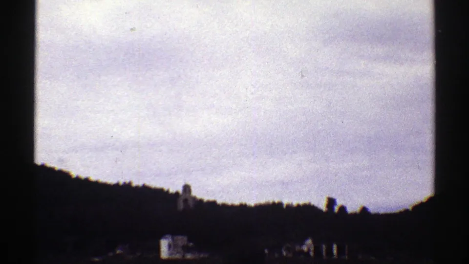 1969 Colorado home-movie frame with a dark hillside, small distant buildings, and a steeple-like structure below a bright sky.