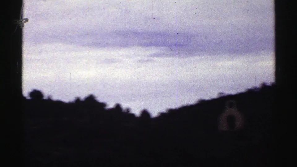 Colorado home-movie frame from 1969 showing clouds above a sloped silhouetted ridge and a small structure at lower right.