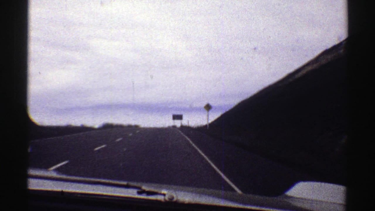 Highway seen through a car windshield, with a dark hillside and roadside signs ahead.