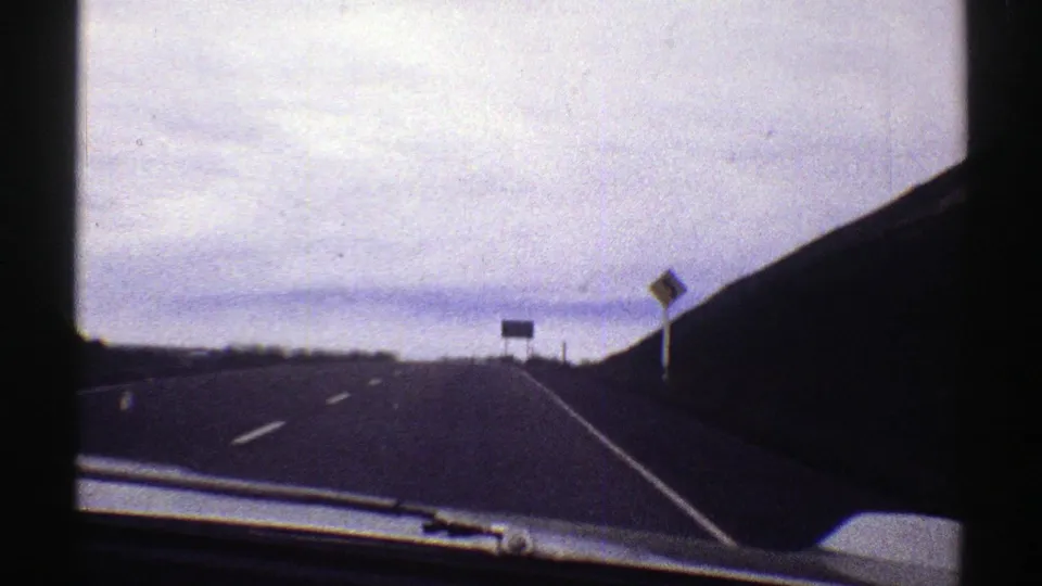 1969 Colorado 8mm frame of a curving mountain highway filmed through a windshield, with signs and overcast sky ahead.