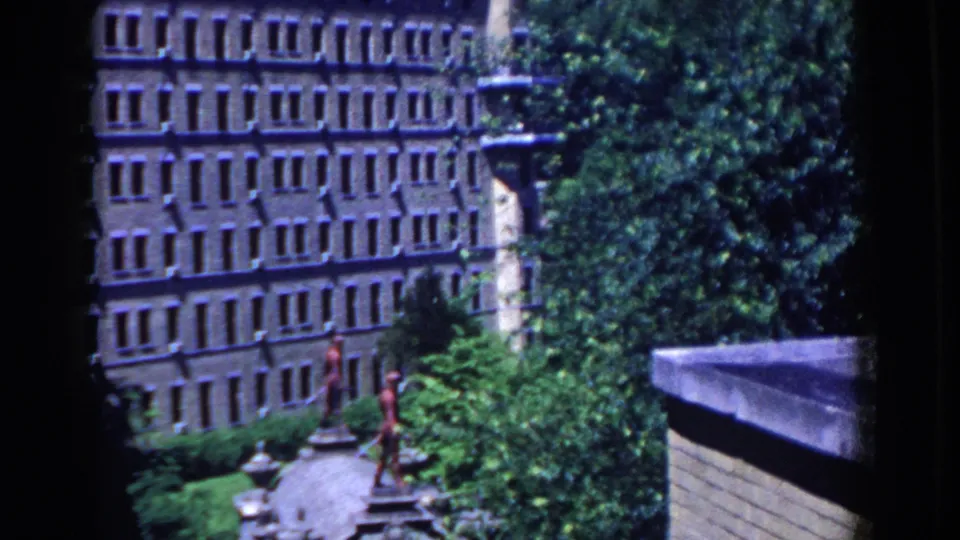 1958 French Lick frame showing a brown institutional-looking building behind trees, with pedestrians on the landscaped path below.