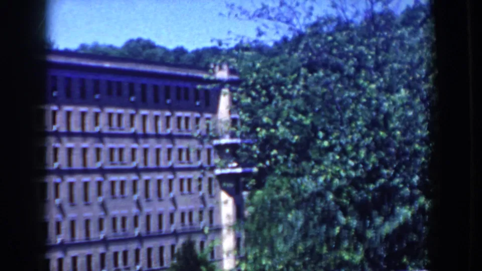 1958 French Lick view of a brown multi-story building partly obscured by dense green trees from an elevated balcony angle.