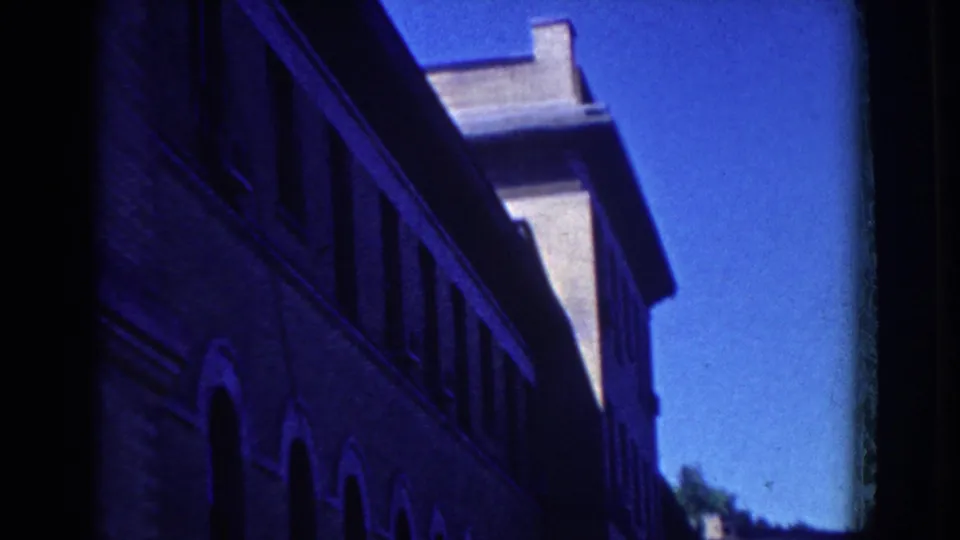 1958 French Lick footage frames a tall brick building in shadow against blue sky, with architectural details partly visible.
