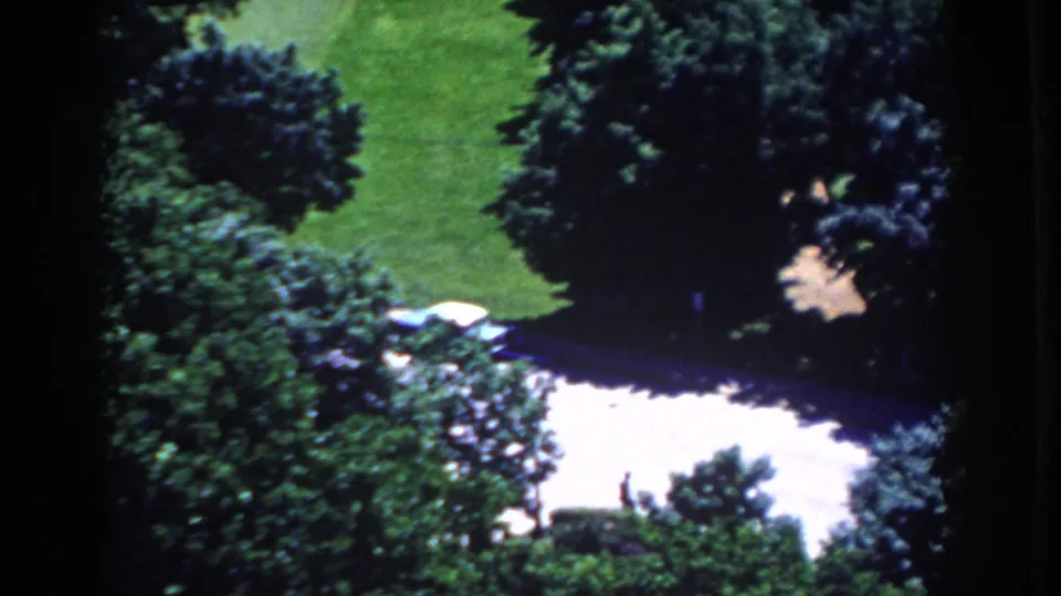 1958 8mm home-movie view in French Lick, Indiana, showing trees, lawn, driveway, and a partly visible parked car.