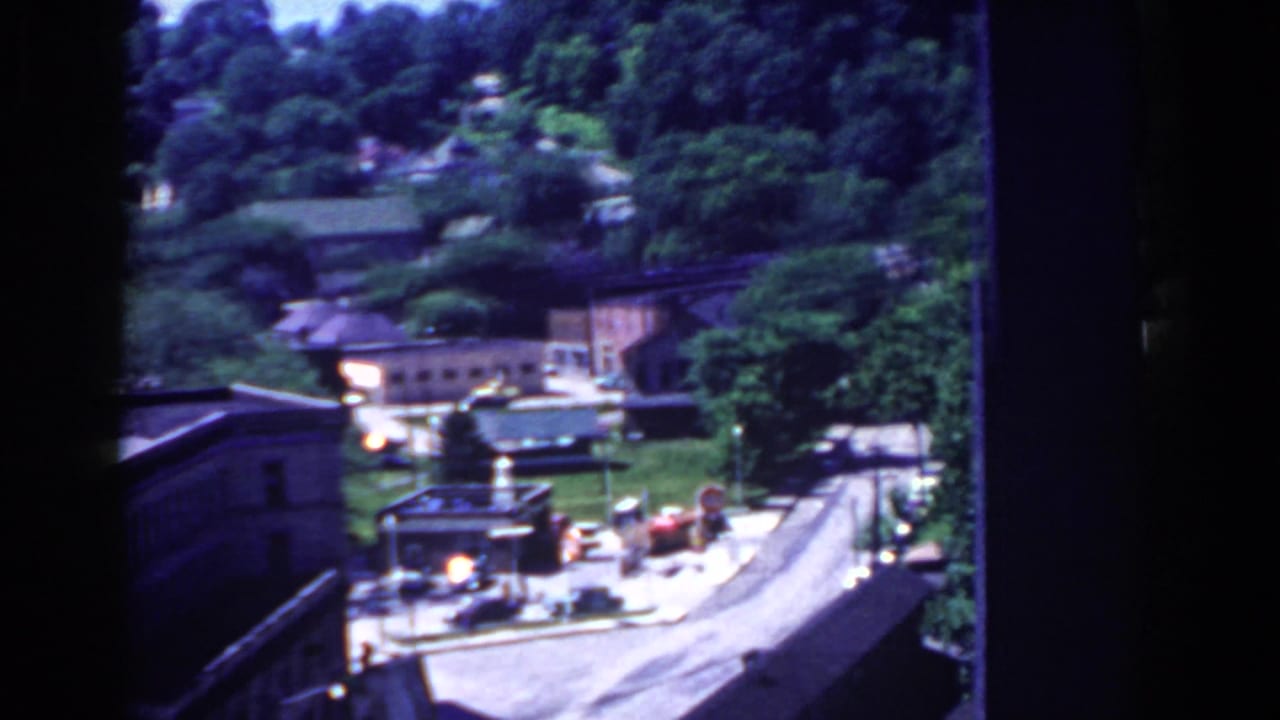 1958 French Lick, Indiana view from above with a curving road, rooftops, dense trees, and distant wooded hills.