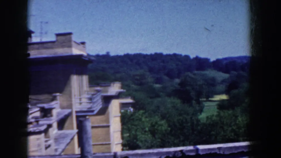 Elevated French Lick view with a roofline in the foreground, dense trees, open green land, and rolling hills under blue sky.