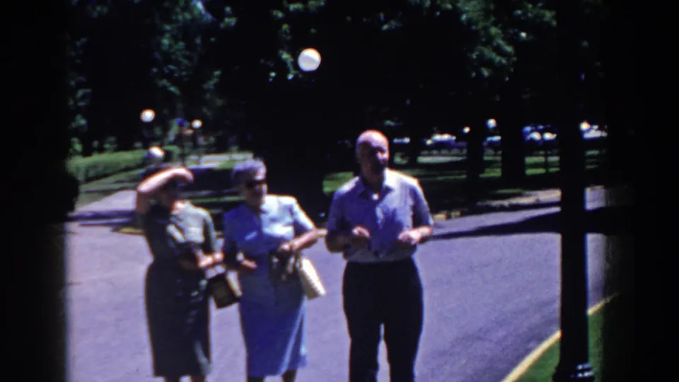 In French Lick, Indiana, three adults walk toward the camera on a shaded paved road, with globe lamps and trees behind them.