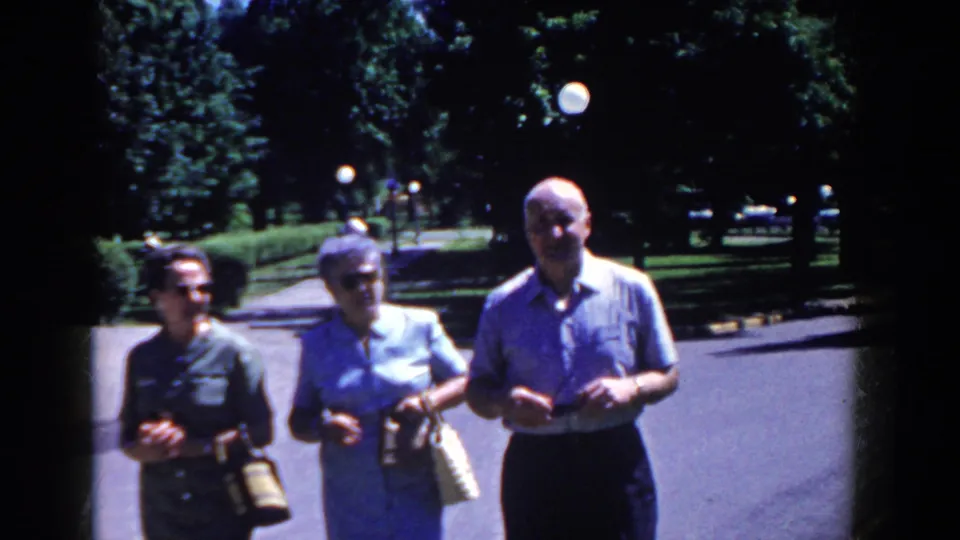 Near the end of the clip, the man and two women are close to the camera on the French Lick road, with trees, lamps, and parked cars in the background.