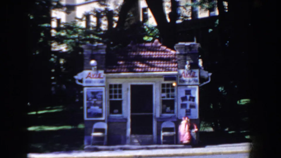 A small French Lick street kiosk or booth appears beneath trees, with signage, display panels, sidewalk, and a building in the background.