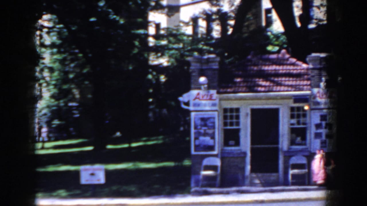 1958 French Lick home-movie frame showing a small kiosk-like building, poster signs, trees, lawn, and a person near the doorway.
