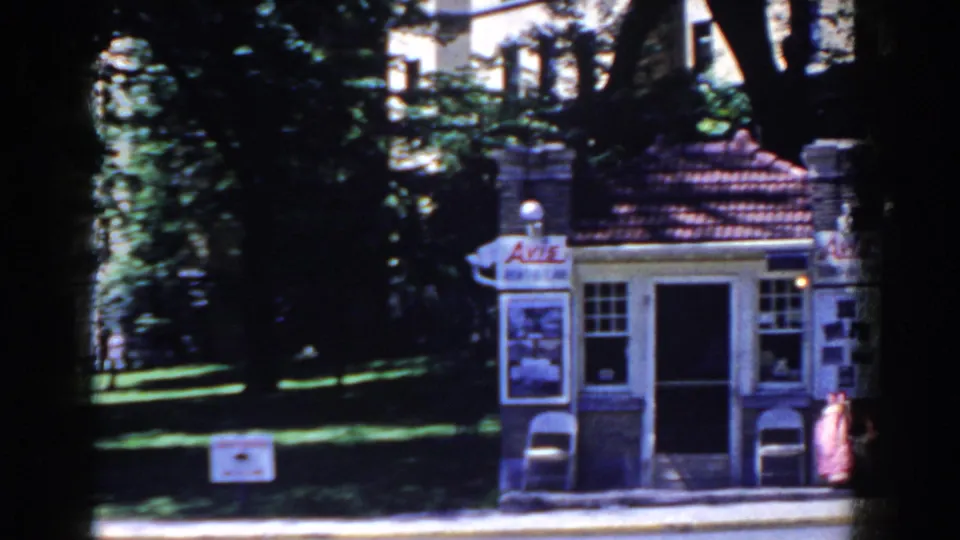 1958 French Lick home-movie frame showing a small kiosk-like building, poster signs, trees, lawn, and a person near the doorway.
