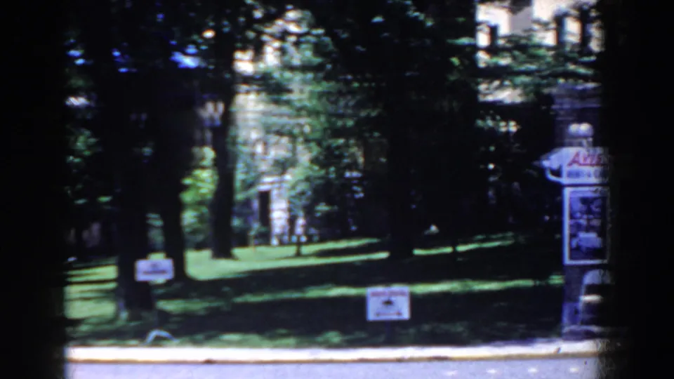 French Lick park view with dark tree shade, green grass, a small sign, globe lamp, and partial commercial-style sign at the right edge.