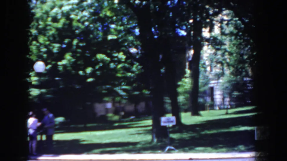 1958 French Lick frame with two people at the left edge, a shaded lawn, mature trees, small sign, and globe lamp near the walkway.
