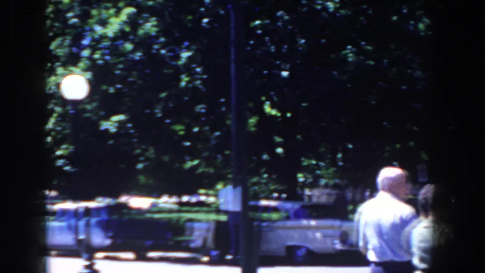 Pedestrians are seen from behind near a tree-lined French Lick street with parked cars and globe streetlamps in 1958 footage.
