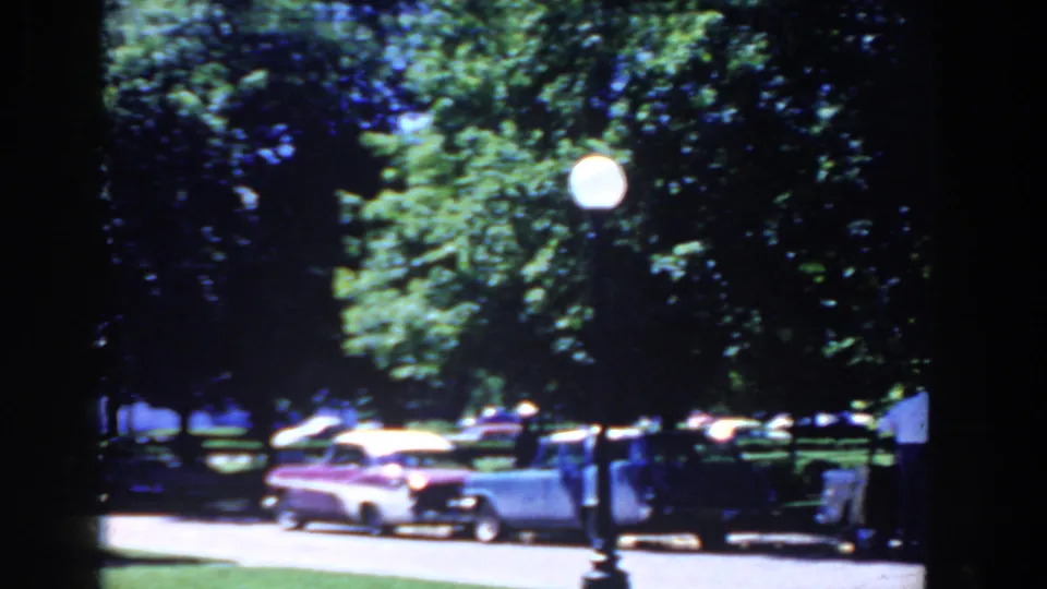 A row of late-1950s cars sits along a sunny French Lick street, with trees, a sidewalk edge, and a small sign visible.