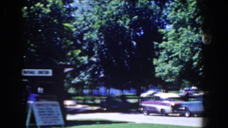 The frame shows parked classic cars, a small roadside sign, and dense trees along a French Lick street in 1958 home-movie footage.