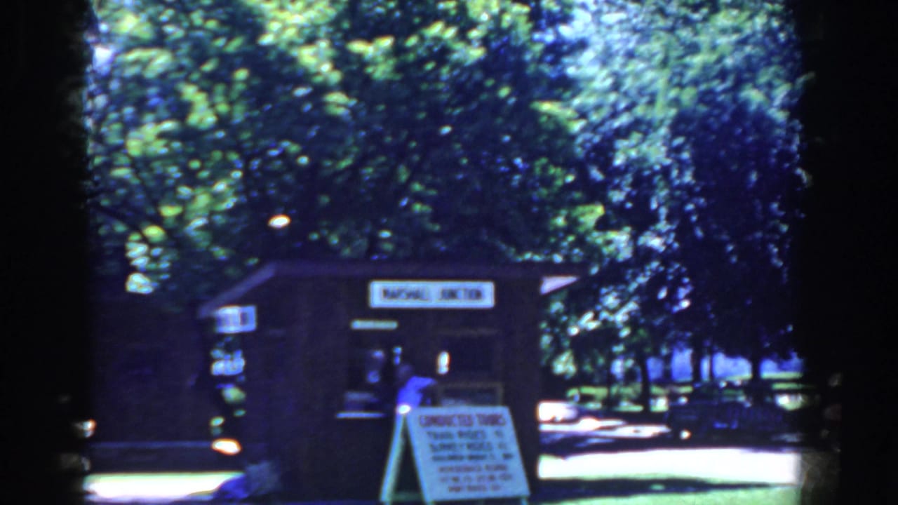 Tour booth and sign in a shaded park with a light-colored parked car.