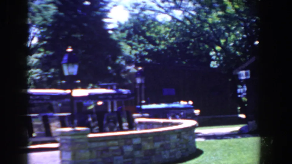 1958 home-movie view in French Lick, Indiana, with a stone fountain, lamppost, shaded booth, trees, and park lawn.
