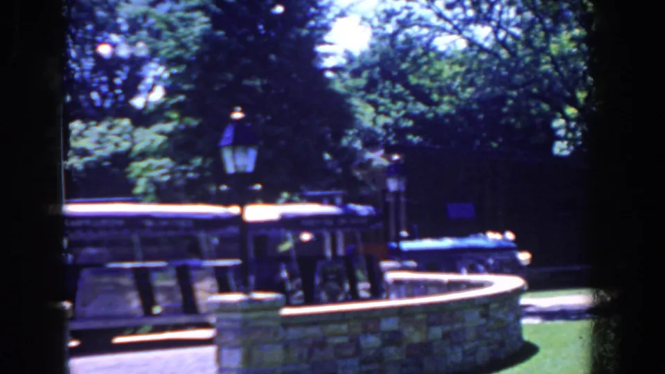 1958 French Lick, Indiana view of a parked open-sided tour bus framed by a stone wall, lampposts, trees, and lawn.