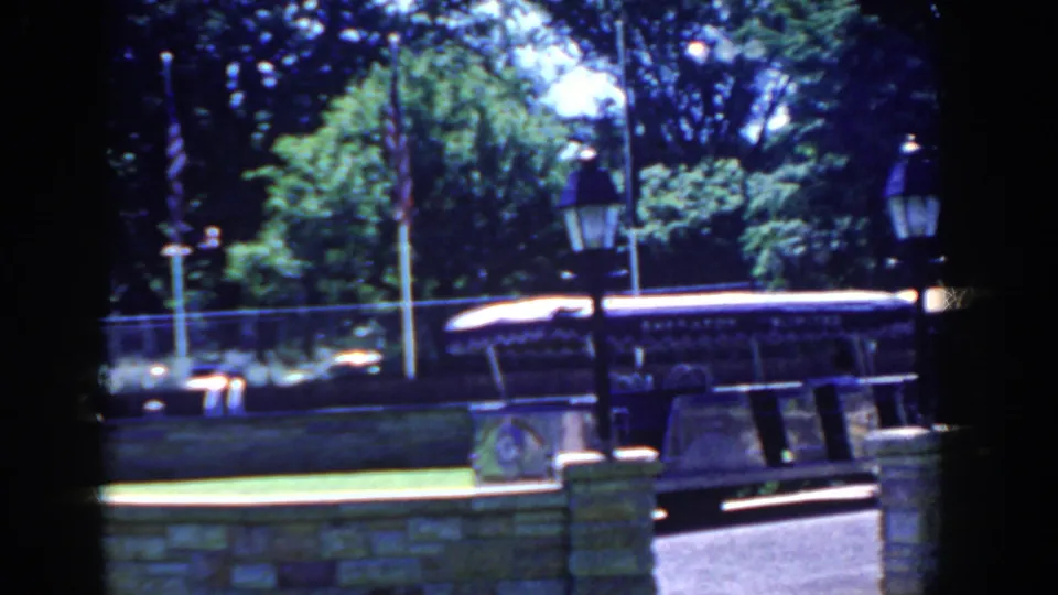 A side view shows the open-sided bus along the wall, with a fence, lawn, flagpole, and leafy trees behind it in French Lick.