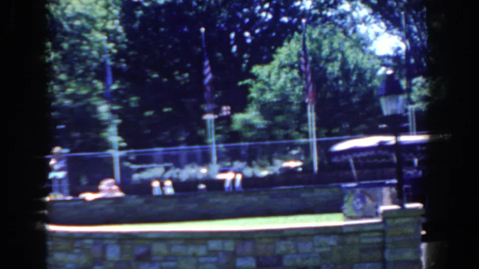 The final frame shows the bus partly cropped at right, with a stone wall, American flag, lamppost, fence, and shaded trees visible.
