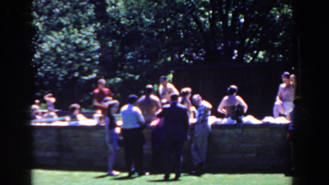 Adults gather on grass beside a low stone pool edge under trees.