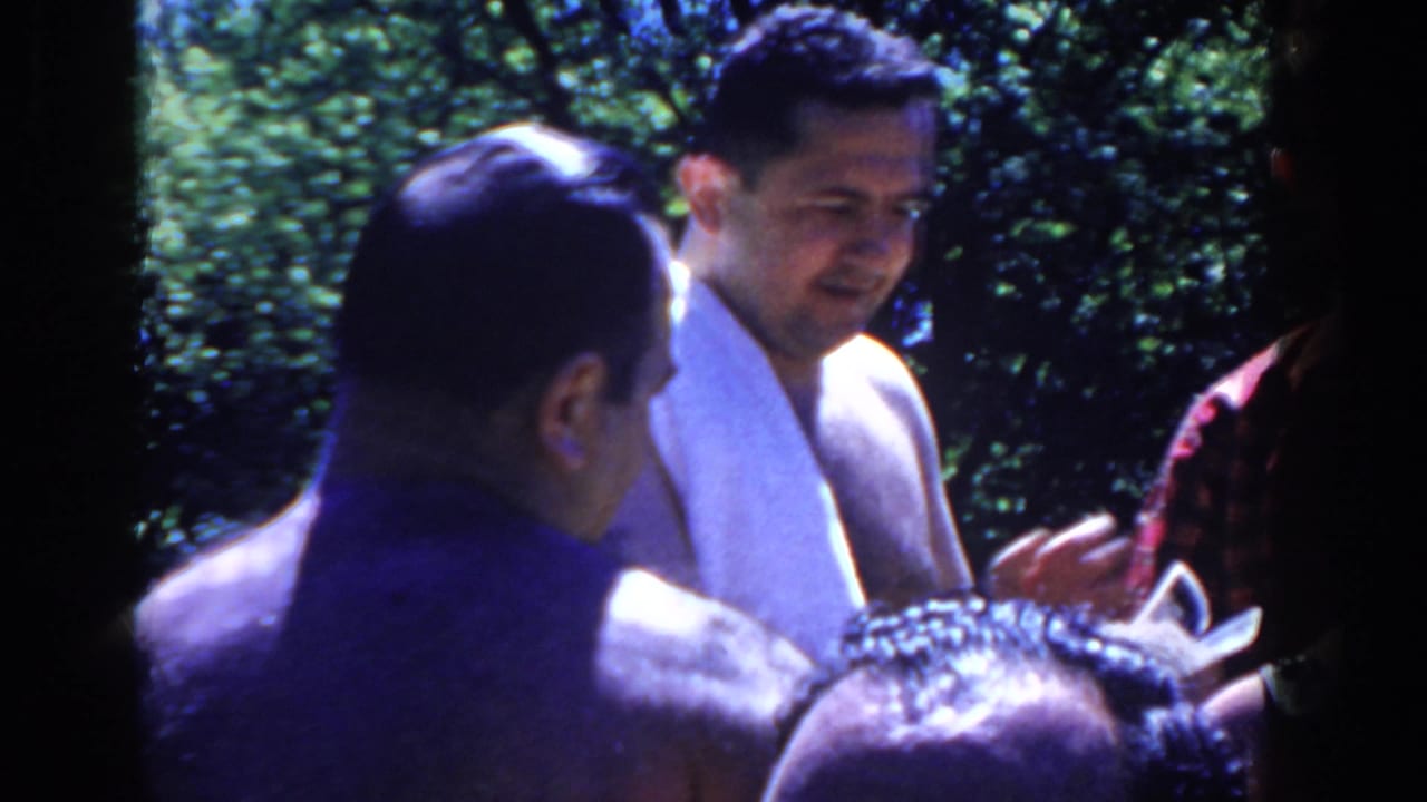 A man with a towel smiles near other poolside adults and greenery.