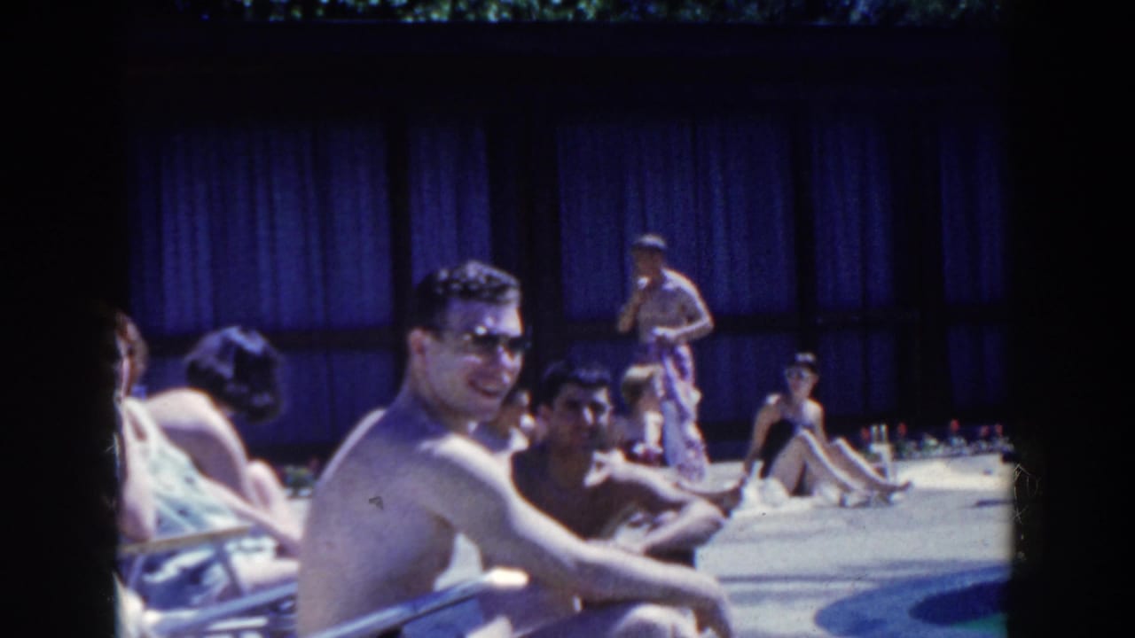 Man in sunglasses sits in a white pool chair beside other people at an outdoor pool area.