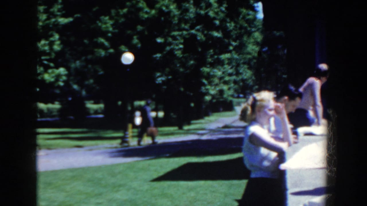 The French Lick resort setting shows poolside visitors beside green lawns, trees, a paved path, and a round lamppost in bright sun.