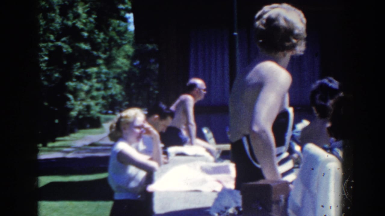 1958 French Lick home-movie frame showing adults in swimsuits gathered beside a pool, with trees and shaded seating nearby.