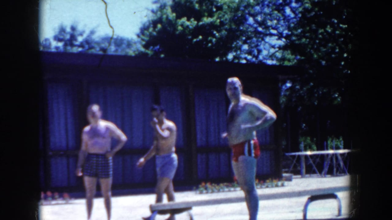 Three men in swim trunks stand on a sunny pool deck near a low building.